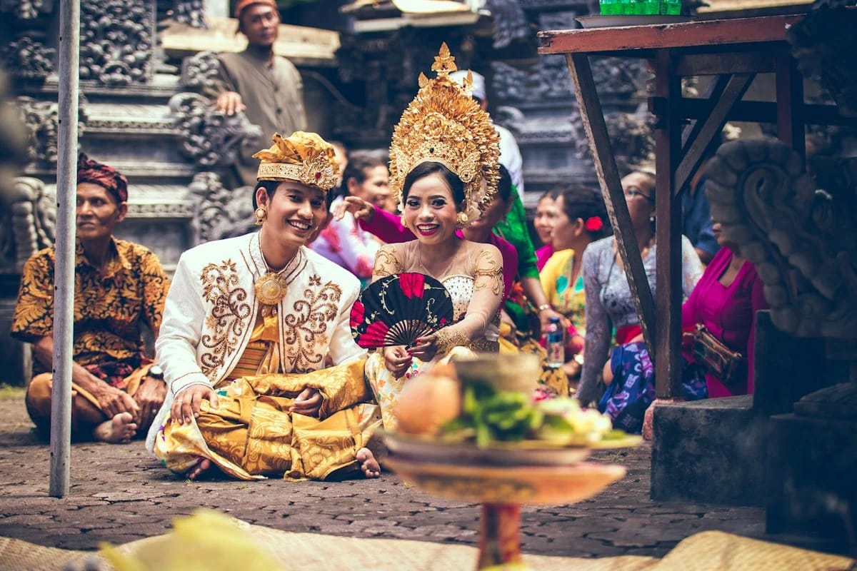 Pengantin Tradisi Jawa duduk berdampingan di halaman joglo, mengenakan busana emas dan mahkota bunga